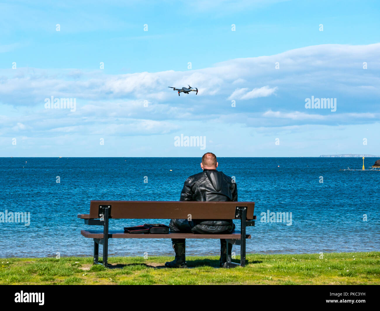 West Bay, North Berwick, East Lothian, Scotland, UK, 12th September 2018. UK Weather: A beautiful sunny but windy day on the Eastern Scottish coastline in the Firth of Forth. A man wearing motorbike leathers sits on a bench and flies a drone Stock Photo