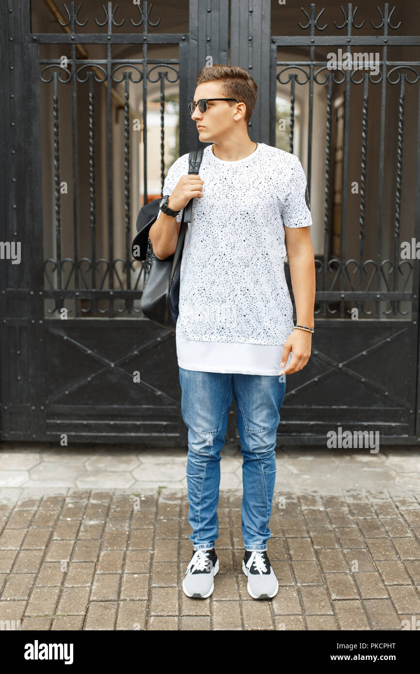 Young handsome hipster man in a white t-shirt, blue jeans and sneakers standing on the street. Backpack with a cap on his shoulder. Stock Photo