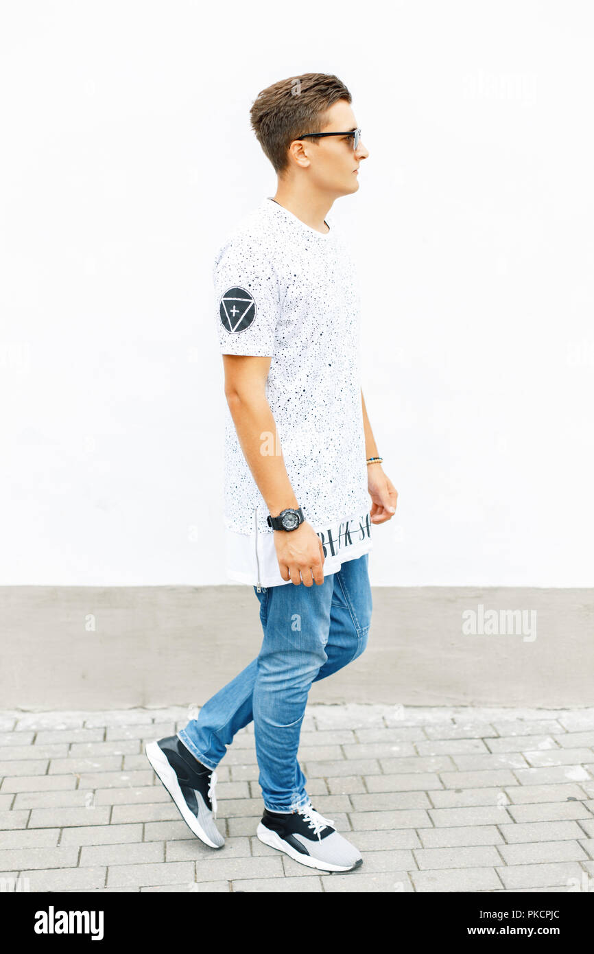 Young handsome man in a white T-shirt, blue jeans and sneakers standing near a white wall. Stock Photo