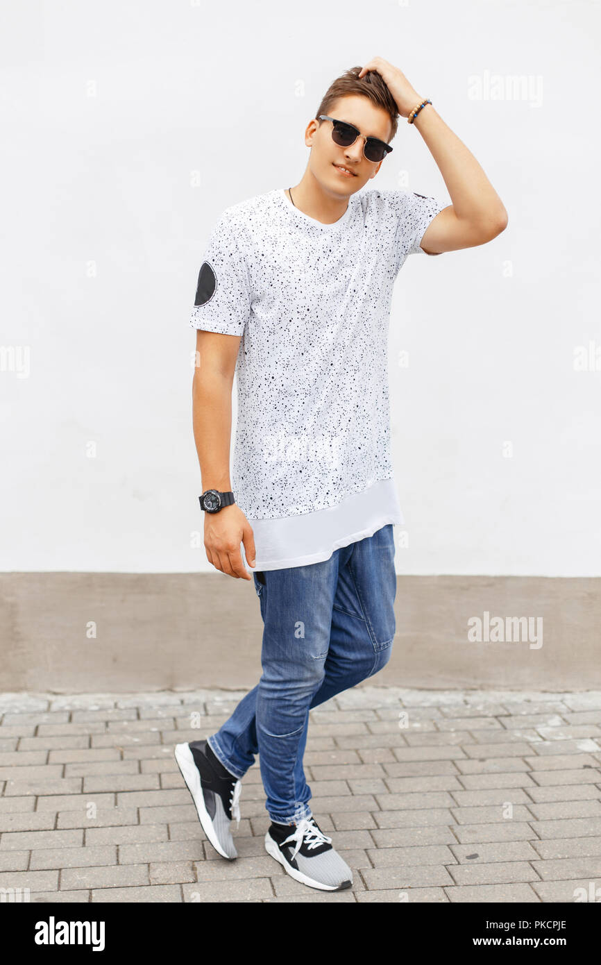 Young handsome man in a white T-shirt, blue jeans and sneakers standing near a white wall. Stock Photo