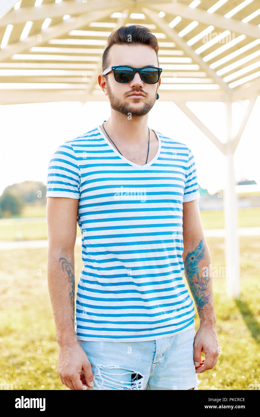 Trendy handsome hipster man with a beard in sunglasses in a trendy shirt stands on the beach. Stock Photo