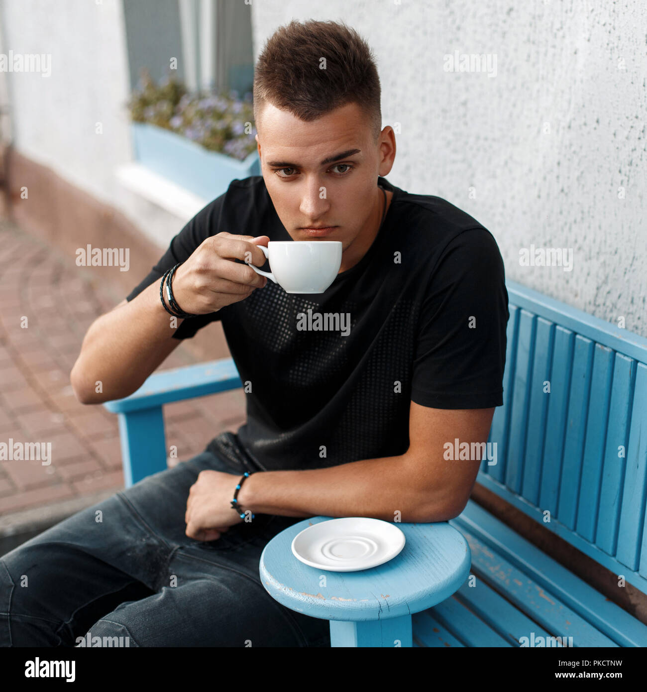 Handsome man with the hairstyle in a black shirt sits on a blue bench and drink coffee. Stock Photo