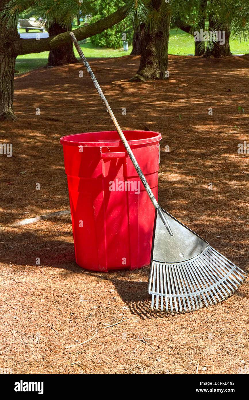 Red plastic trash can and garden rake leaning against it during a