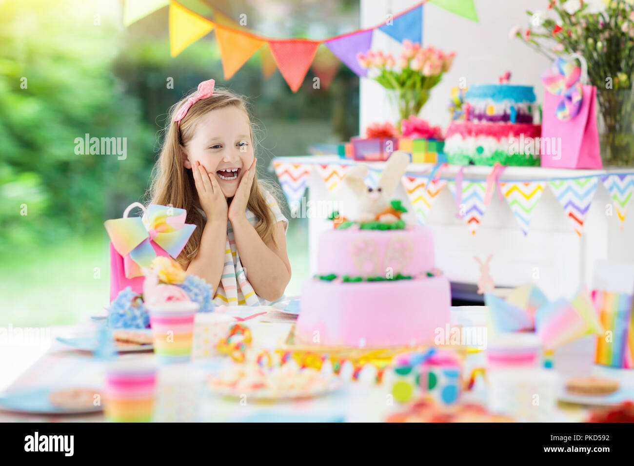 Kids birthday party with colorful rainbow pastel decoration and bunny layer cake. Little girl with sweets, candy and fruit. Balloons and banner at fes Stock Photo