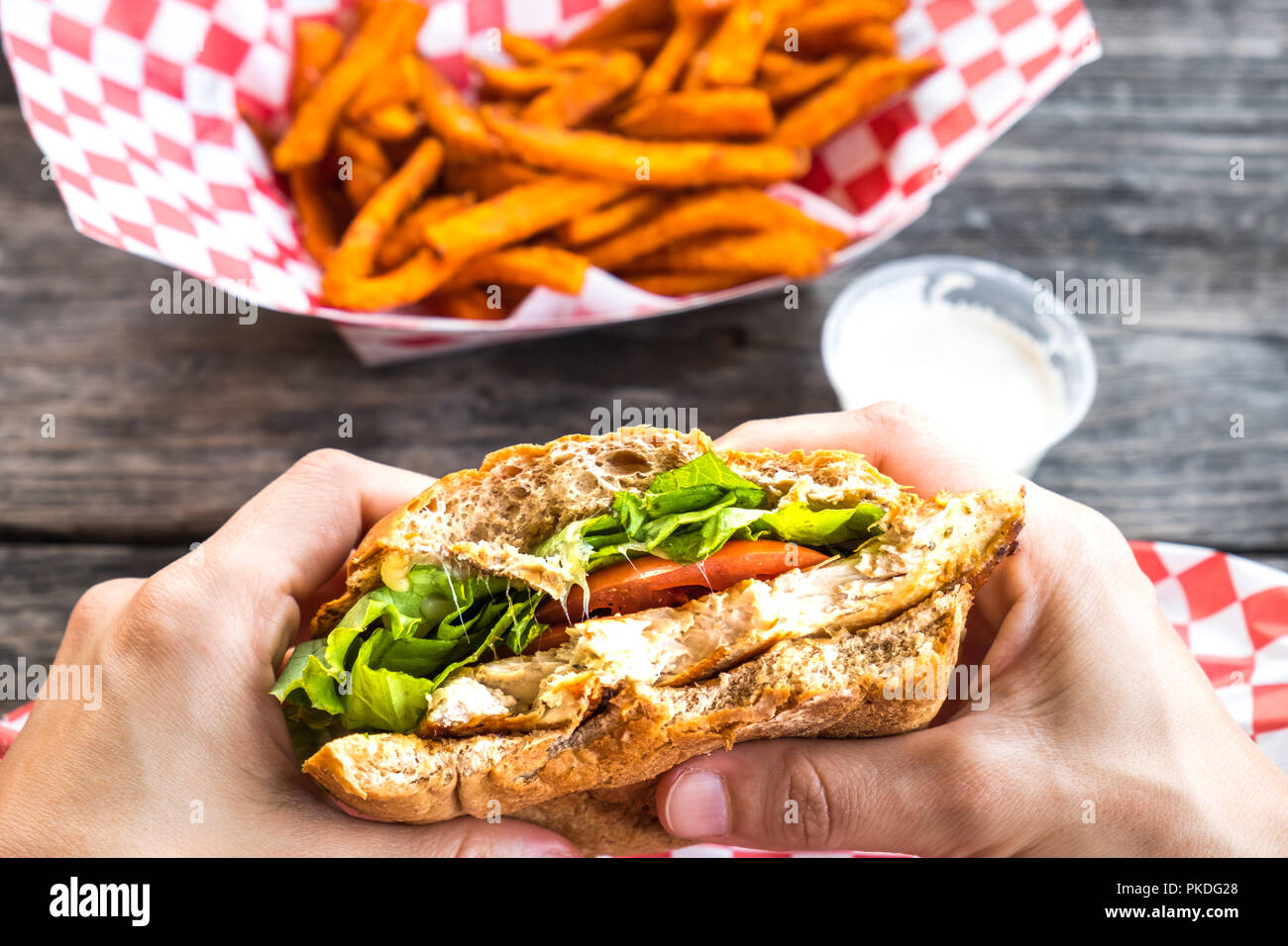 Woman hands hold burger style grilled chicken breast sandwich with lettuce and tomatoes and a side of sweet potato fries Stock Photo