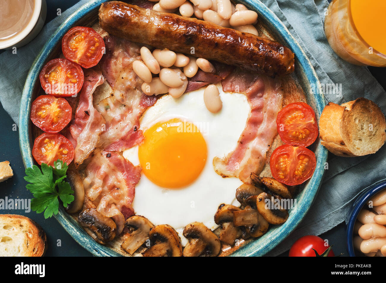 Traditional English breakfast close-up on a blue background, top view. Fried egg with sausage, mushrooms, beans, tomatoes and bacon. Stock Photo