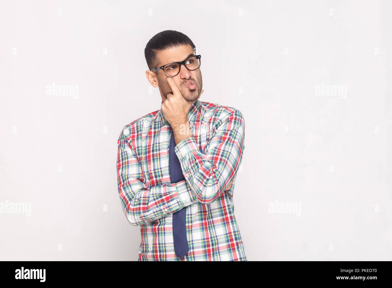Portrait of confused handsome bearded businessman in colorful checkered shirt, blue tie and eyeglasses standing touching face and looking away. indoor Stock Photo