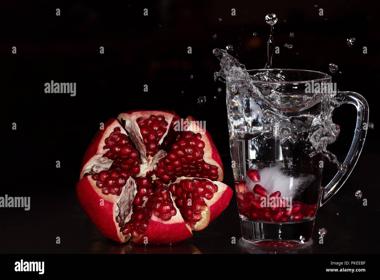 A splash of pomegranate resting in a glass with ice and raw opened fruit displayed on the left Stock Photo