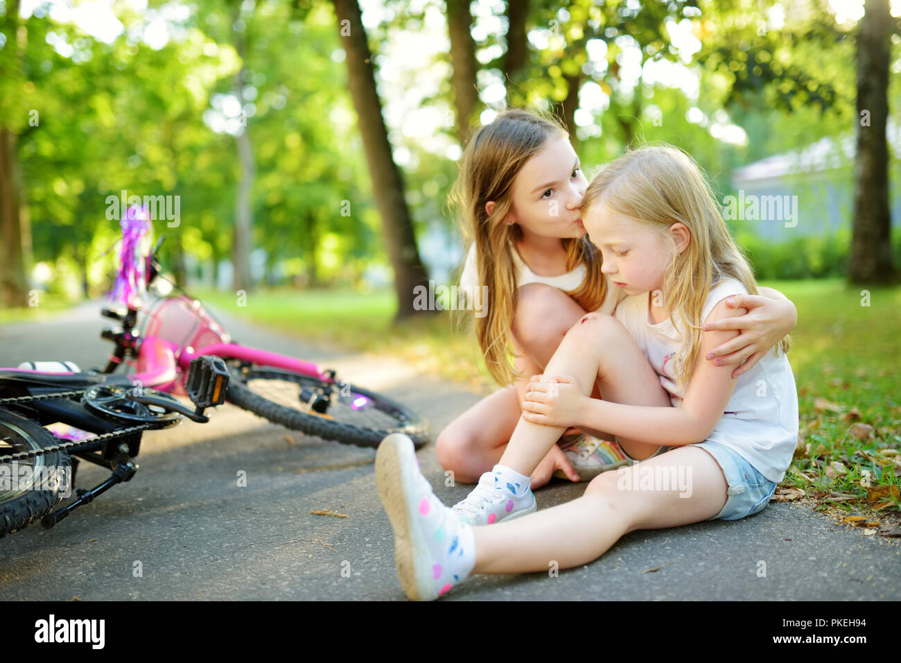 Adorable girl comforting her little sister after she fell off her bike at summer park. Child getting hurt while riding a bicycle. Active family leisur Stock Photo