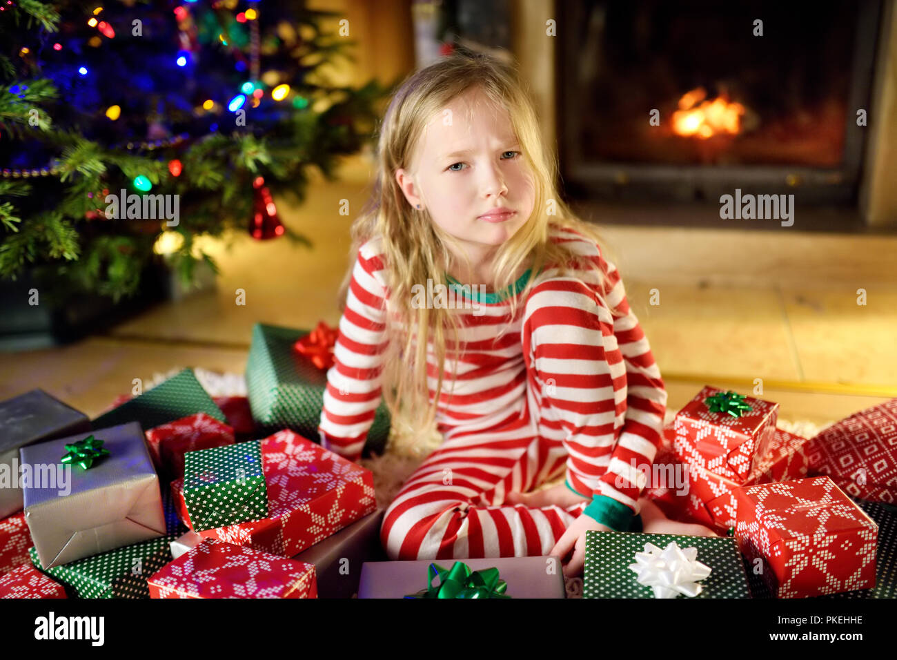 Cute little girl feeling unhappy with her Christmas gifts. Child sitting by a fireplace in a cozy dark living room on Xmas eve. Too many presents for  Stock Photo