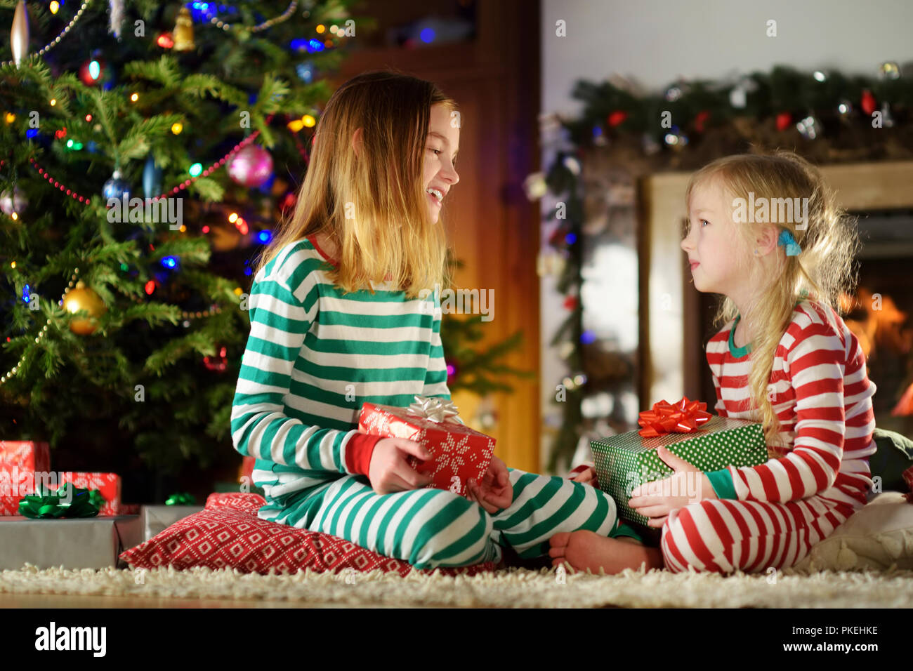 Happy little sisters wearing Christmas pajamas playing by a fireplace in a cozy dark living room on Christmas eve. Celebrating Xmas at home. Stock Photo