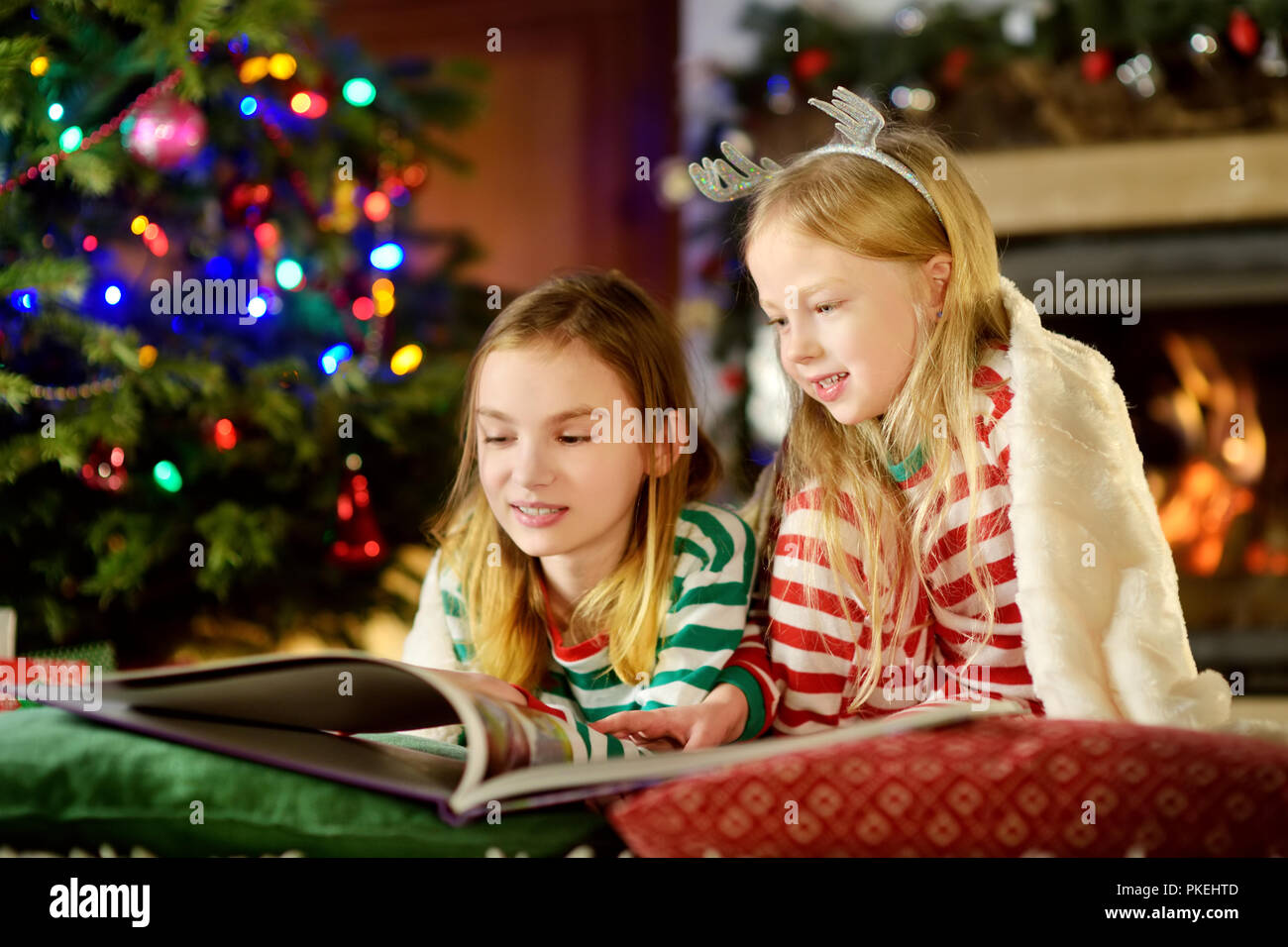 Happy little sisters reading a story book together by a fireplace in a cozy dark living room on Christmas eve. Celebrating Xmas at home. Stock Photo