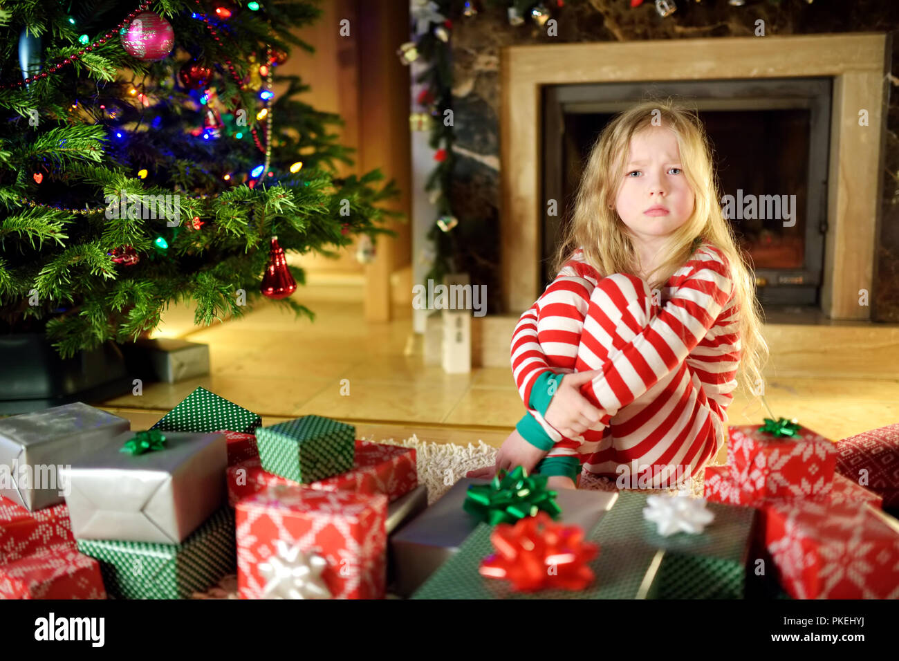 Cute little girl feeling unhappy with her Christmas gifts. Child sitting by a fireplace in a cozy dark living room on Xmas eve. Too many presents for  Stock Photo
