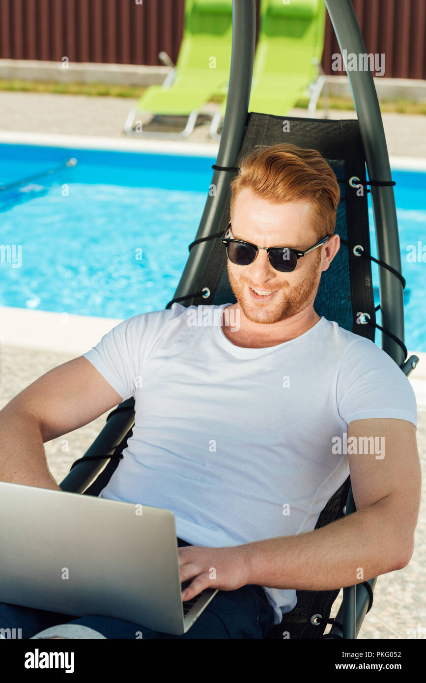 handsome young man sitting in sun lounger in front of swimming pool and working with laptop Stock Photo