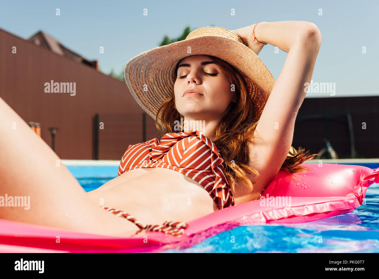 attractive young woman in straw hat and bikini  floating on inflatable mattress at swimming pool Stock Photo