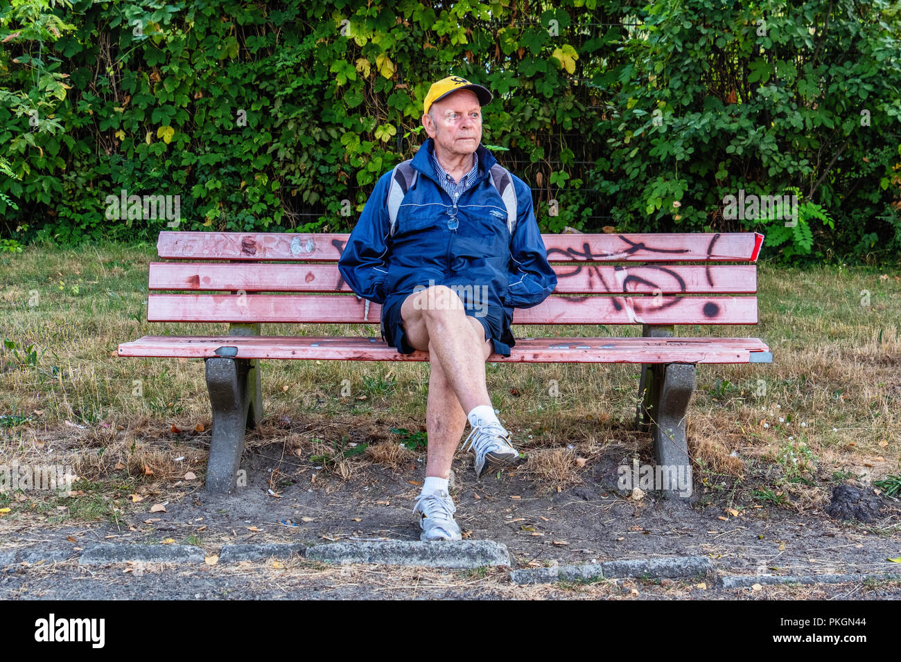 Berlin,Dahlem, Senior man sitting on graffiti covered old bench looking worried. Elderly man wearing shorts & cap sits on bench Stock Photo