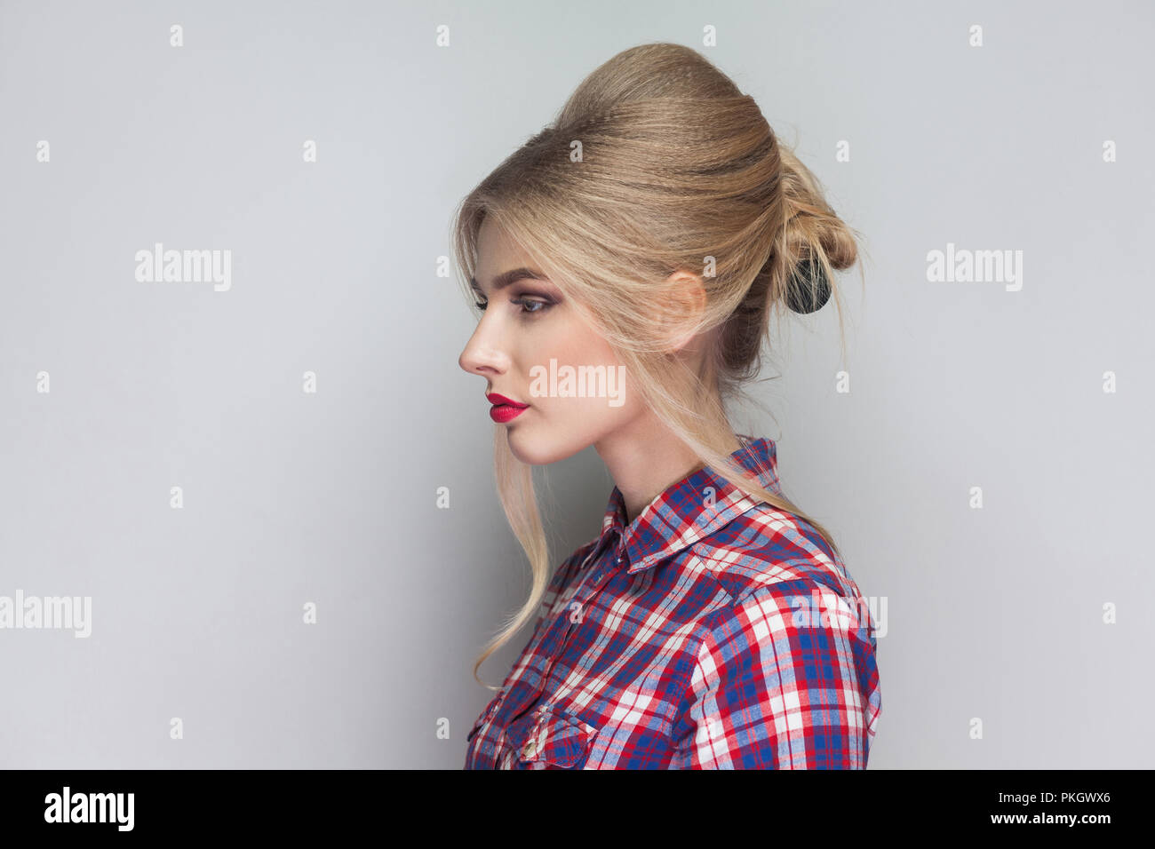 Profile side view of beautiful girl with pink checkered shirt, collected updo hairstyle, red lips and makeup standing with seious face and looking awa Stock Photo
