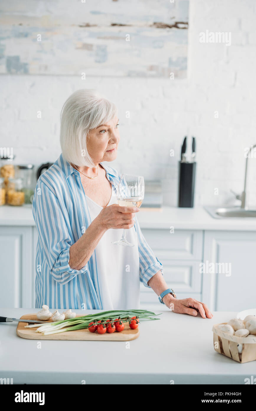pensive senior lady with glass of wine standing at counter with fresh vegetables on cutting board in kitchen Stock Photo
