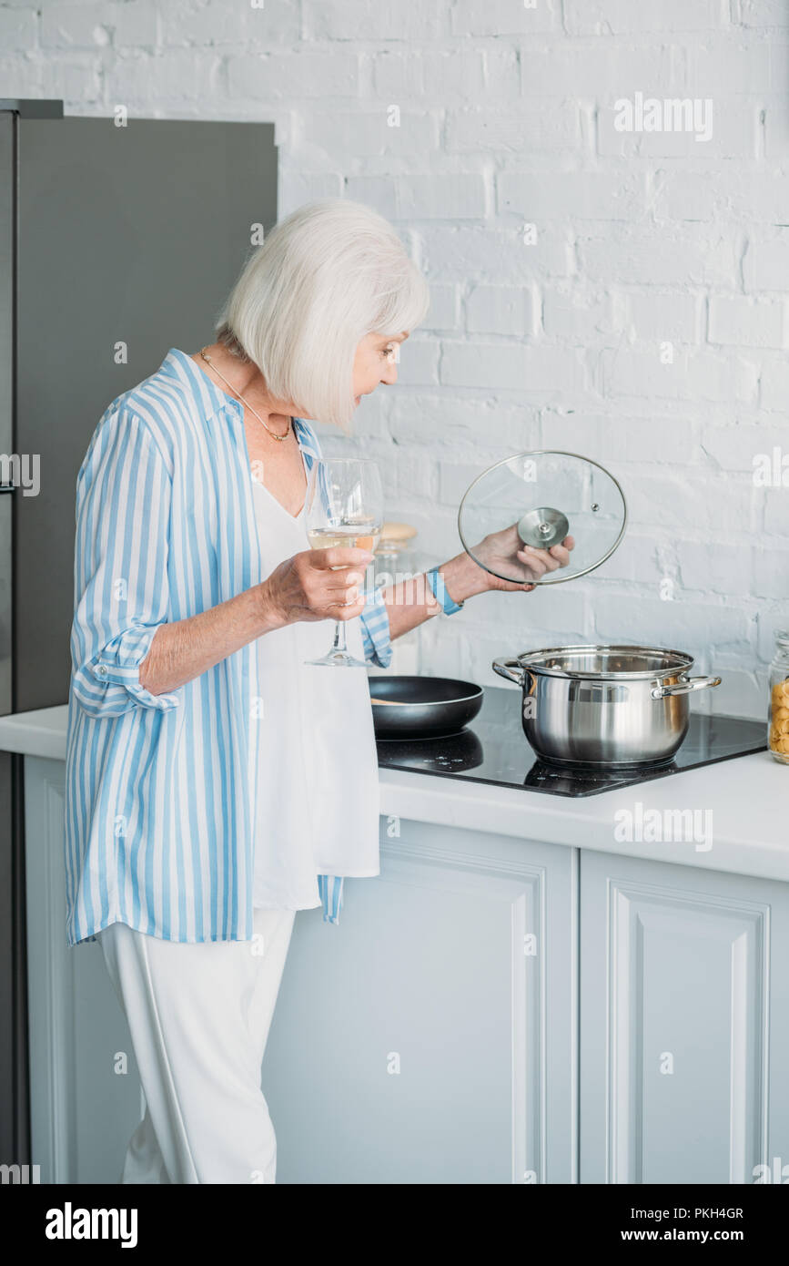 side view of senior lady with glass of wine checking saucepan on stove in kitchen Stock Photo