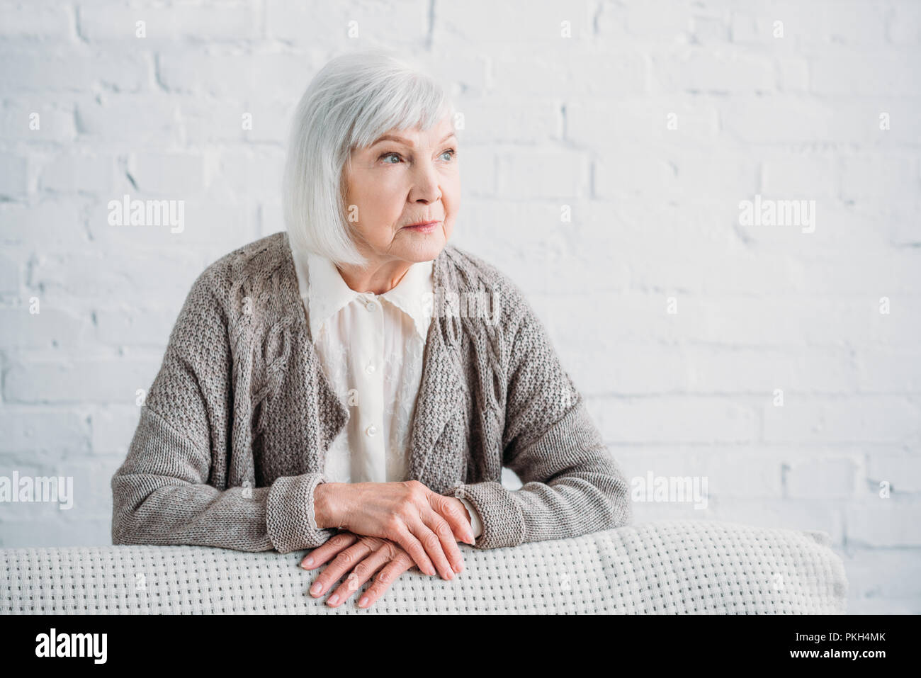 portrait of grey hair lady in knitted jacket leaning on couch and looking away at home Stock Photo