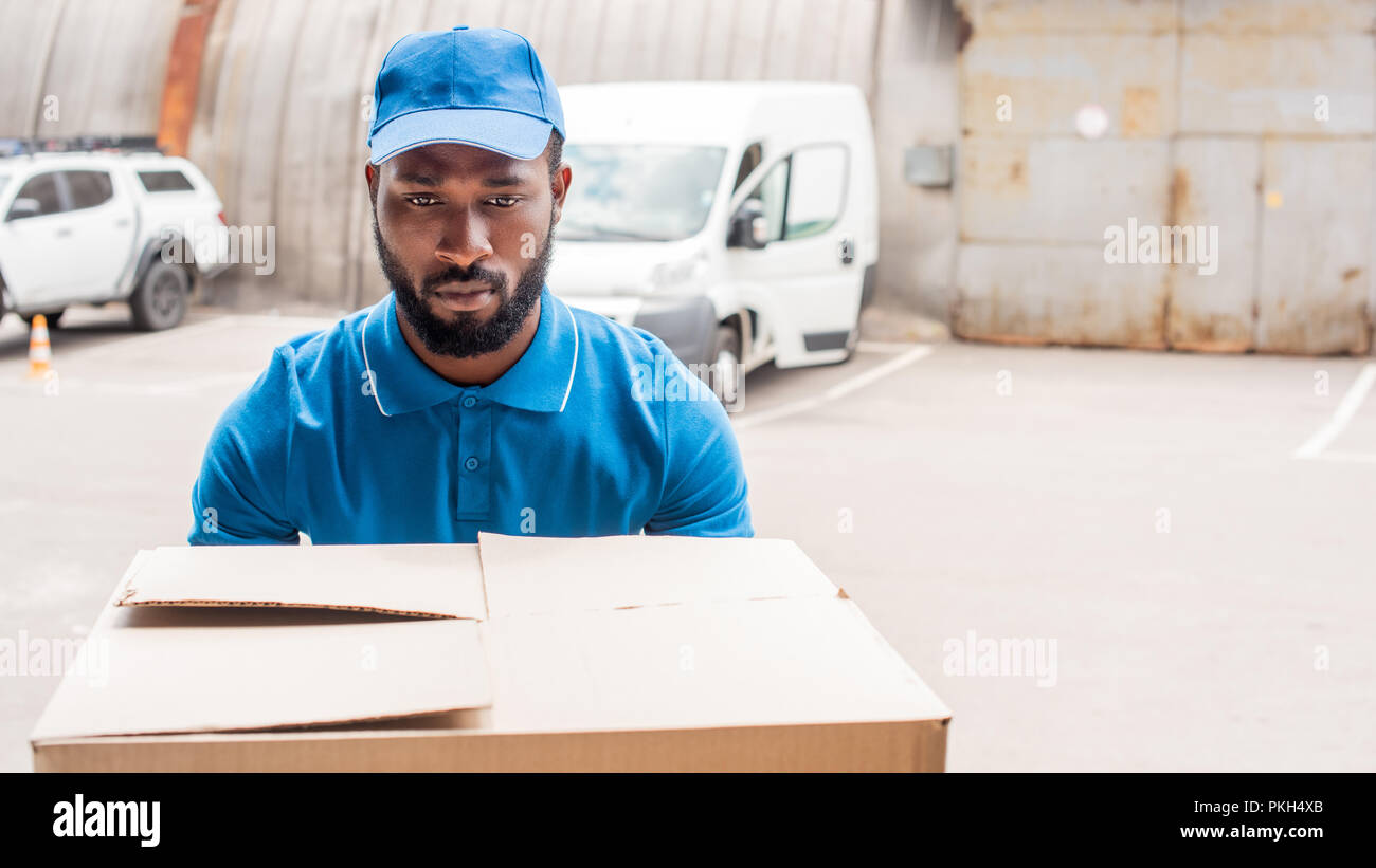 african american delivery man carrying big box with cars on background Stock Photo