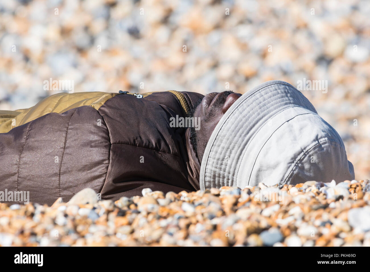 Black man laying on a beach on a warm sunny day in Spring wearing a hat, coat and sunglasses, in the UK. Stock Photo