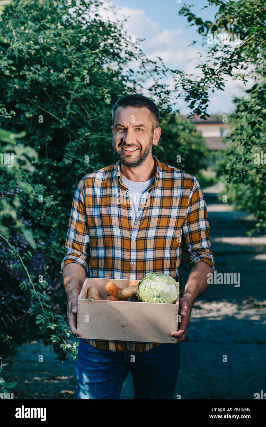 happy farmer holding box with fresh vegetables and smiling at camera ...