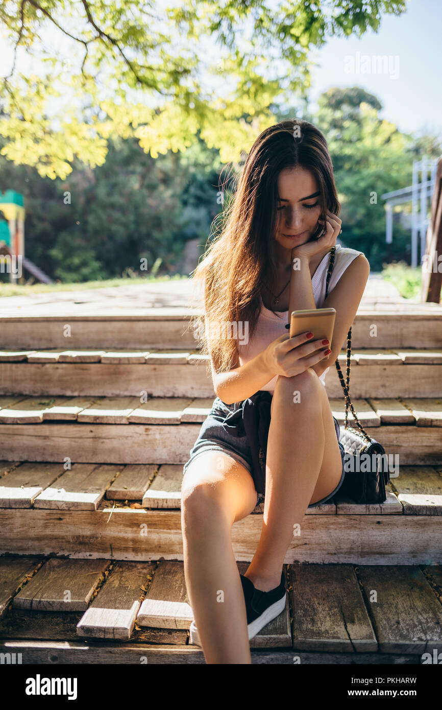 Girl sitting on wooden bench outside in a green park looking at mobile  phone. Young woman wearing shorts and t-shirt is resting and using smart  device Stock Photo - Alamy