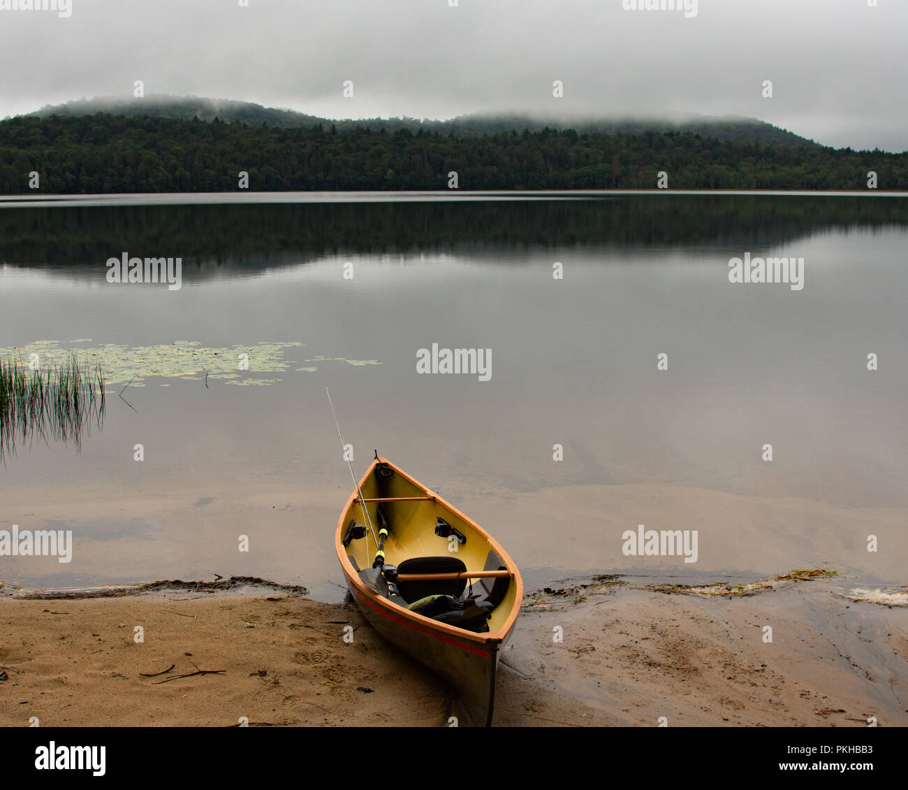 A lightweight canoe with fly fishing rod. paddle and life preserver on a sandy beach on an Adirondack Mountains, NY wilderness lake on a misty morning Stock Photo