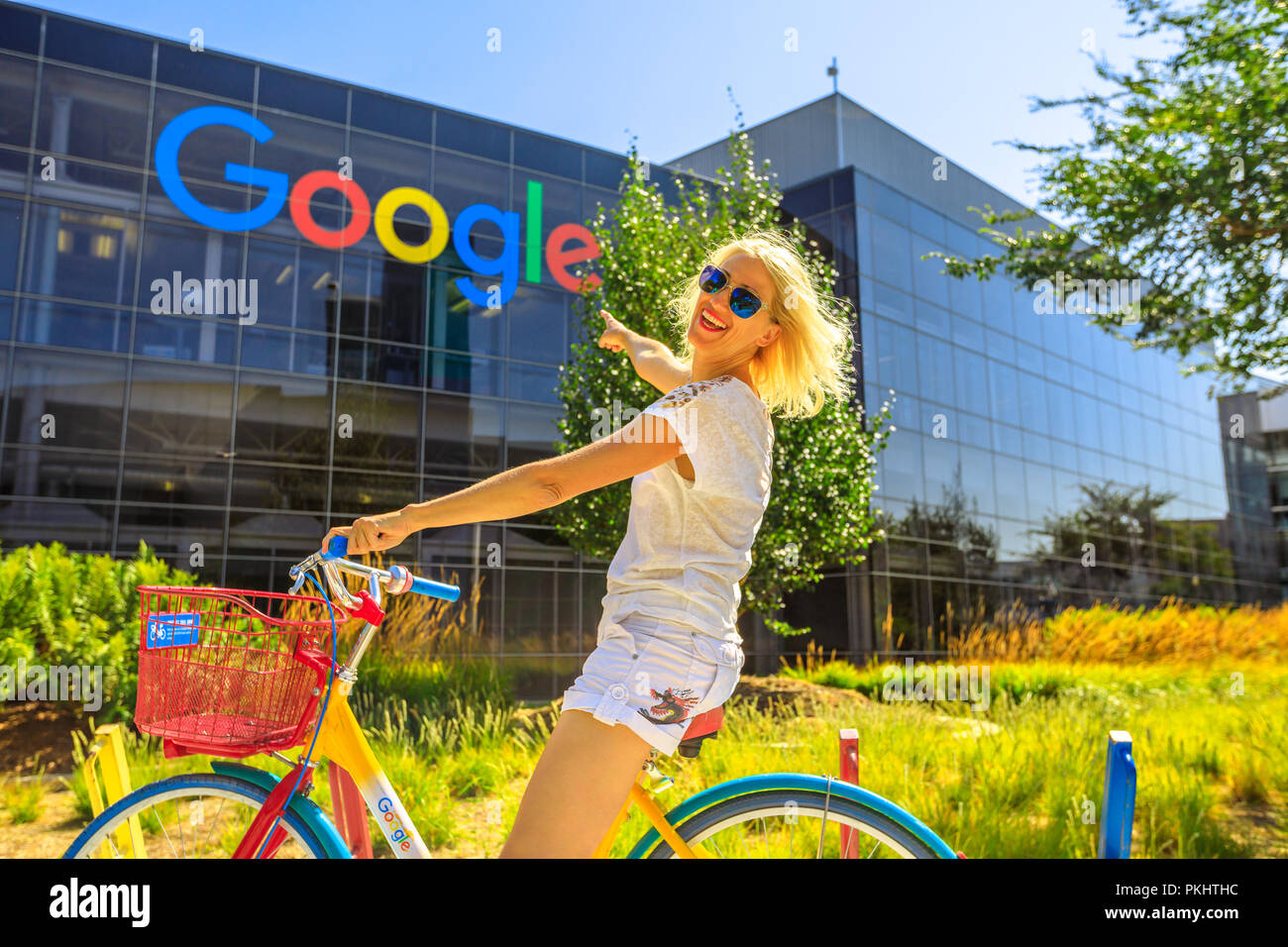 Mountain View, California, USA - August 13, 2018: Smiling woman on Google bike pointing Google sign at Googleplex Headquarters building. A young girl visiting the web leader company in Silicon Valley Stock Photo