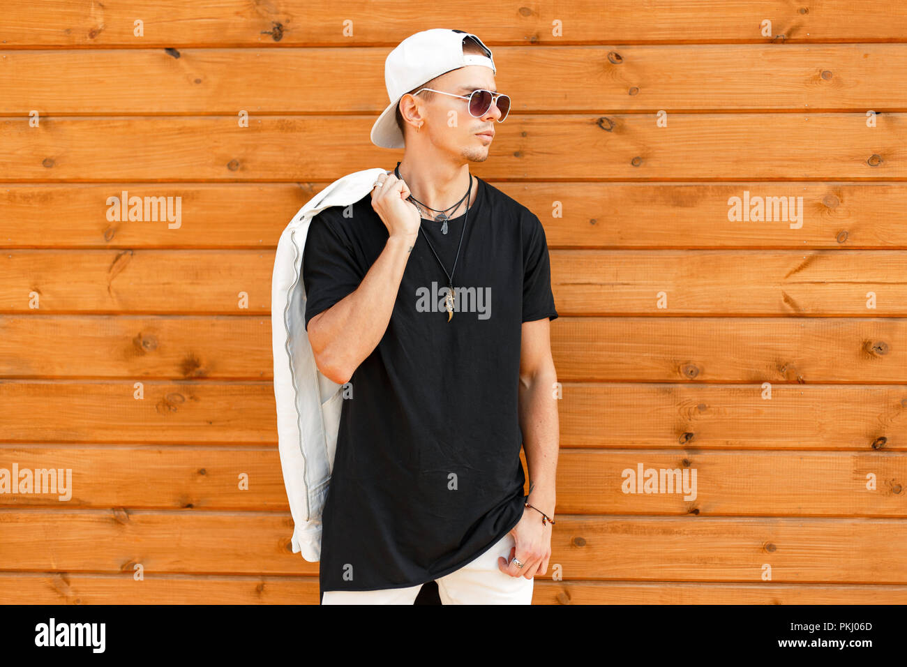handsome fashionable man with sunglasses in a white baseball cap with a  black t-shirt and a white jacket near a wooden wall Stock Photo - Alamy