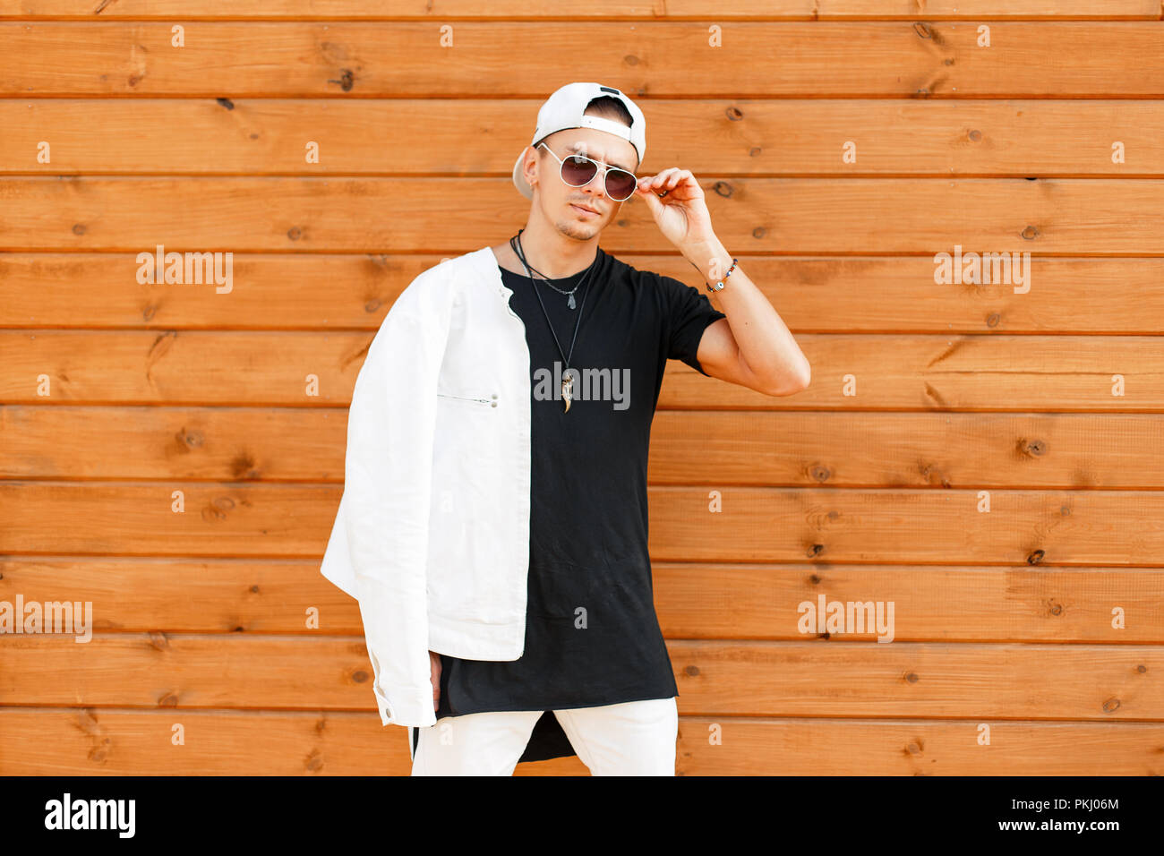 Young stylish man with sunglasses in a white jacket and a T-shirt baseball cap near the wooden wall Stock - Alamy