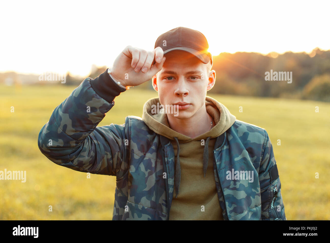 Fashionable young man with a cap in an army jacket with hood in the Park at sunset Stock Photo