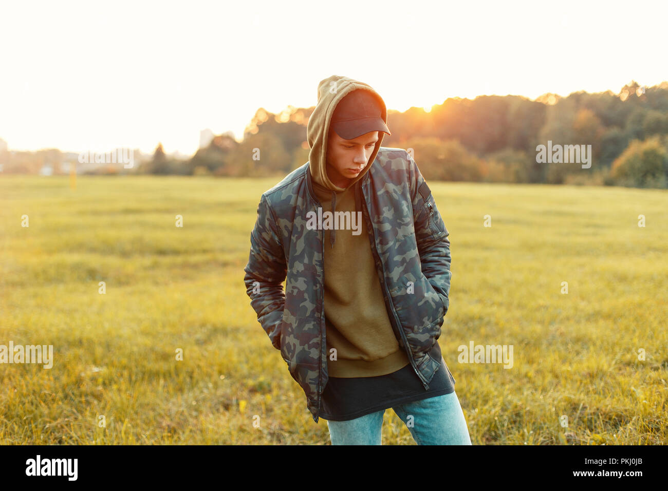 Handsome man with a cap in a military jacket with a hoodie on the nature at sunset Stock Photo