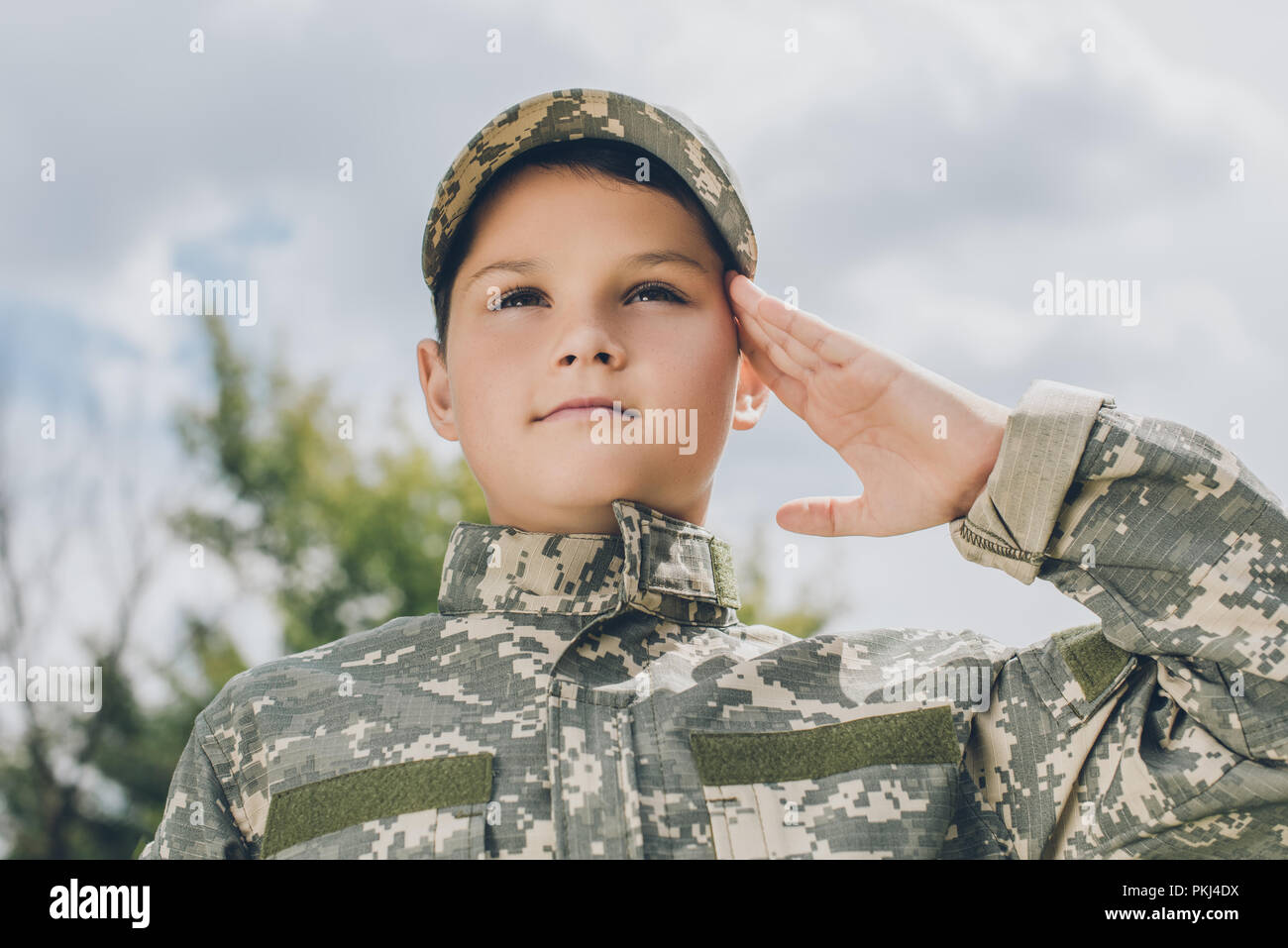 portrait of little boy in camouflage clothing saluting with cloudy sky
