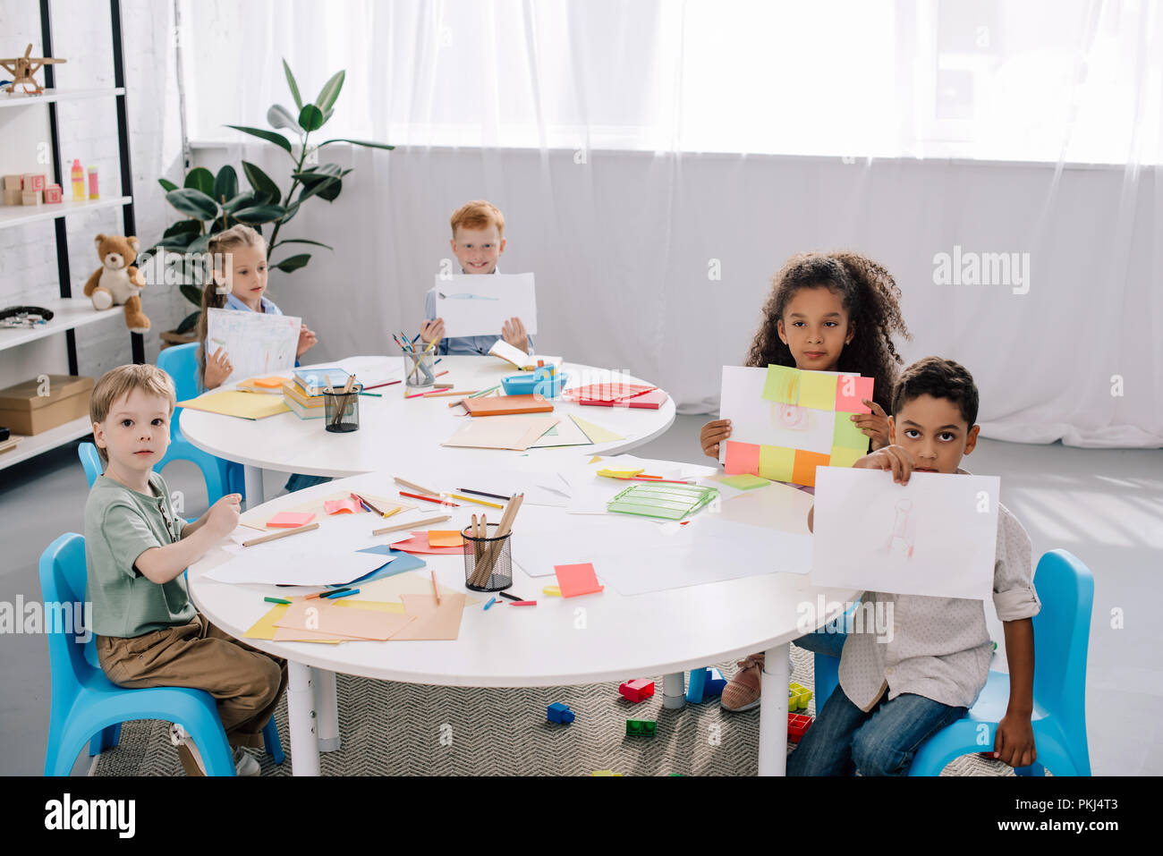 multiethnic kids showing pictures while sitting at table in classroom