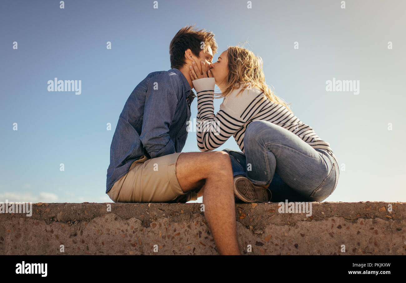 Woman kissing her boyfriend sitting on a sea wall on a sunny day. Couple on vacation in romantic mood sitting outdoors. Stock Photo