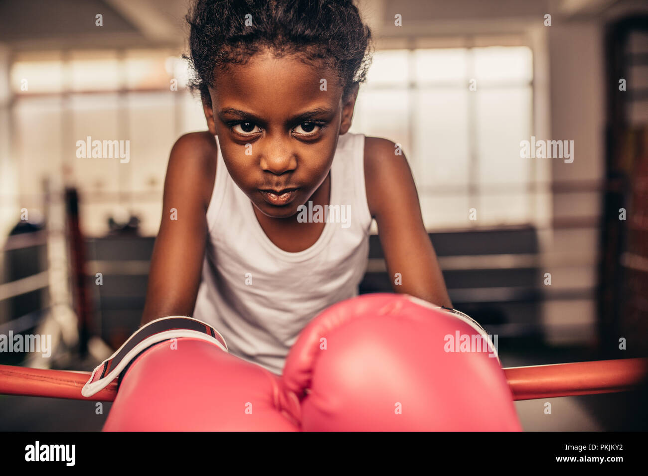 Kid wearing boxing gloves standing inside a boxing ring. Boxer kid resting her hands on boxing ring with determination and focus in her eyes. Stock Photo