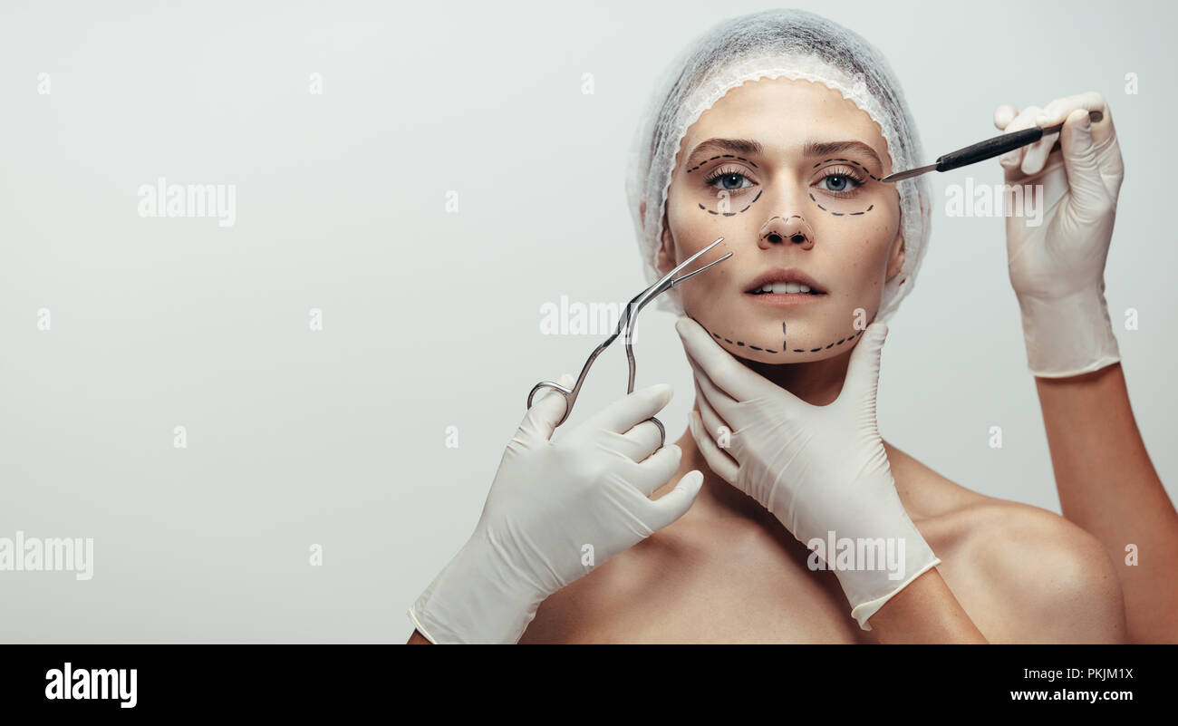 Horizontal shot of woman under going a face lift surgery. Female face with dotted line drawn and beautician holding surgical instruments on grey backg Stock Photo