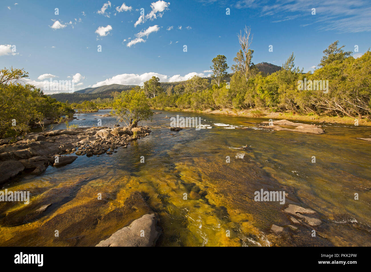 Stunning Australian landscape with rock strewn blue waters of Mann River hemmed by forests at foot of ranges under blue sky with flecks of cloud - NSW Stock Photo