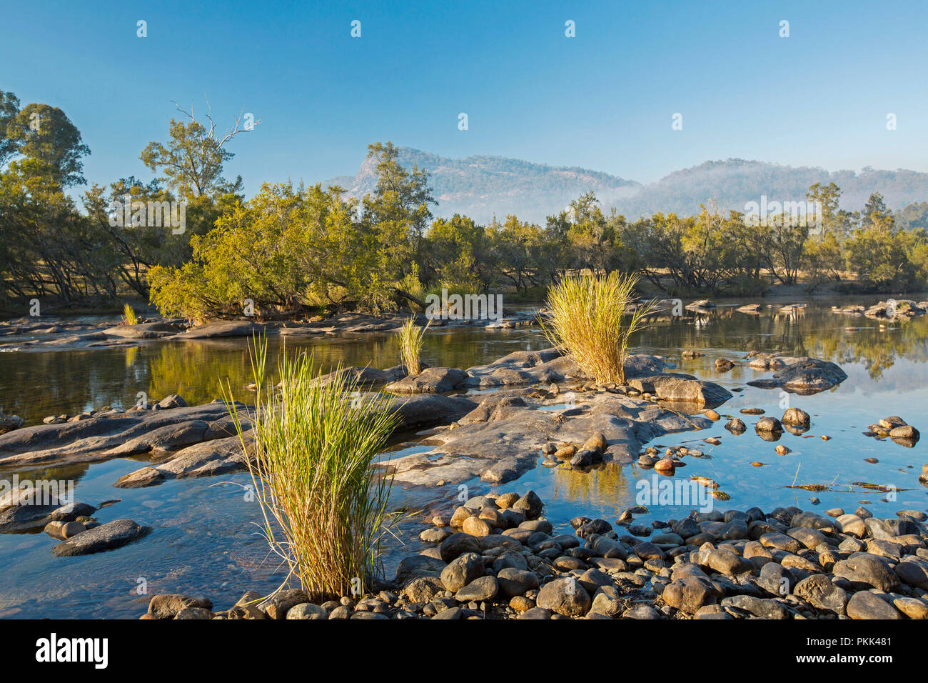 Stunning Australian landscape with rock strewn blue waters of Mann River hemmed by forests at foot of ranges under blue sky of early morning in NSW Stock Photo