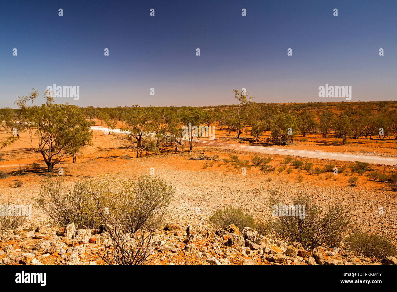 View from stony hill of Australian outback landscape with road slicing across plains, dotted with trees, that stretch to horizon under blue sky in Qld Stock Photo