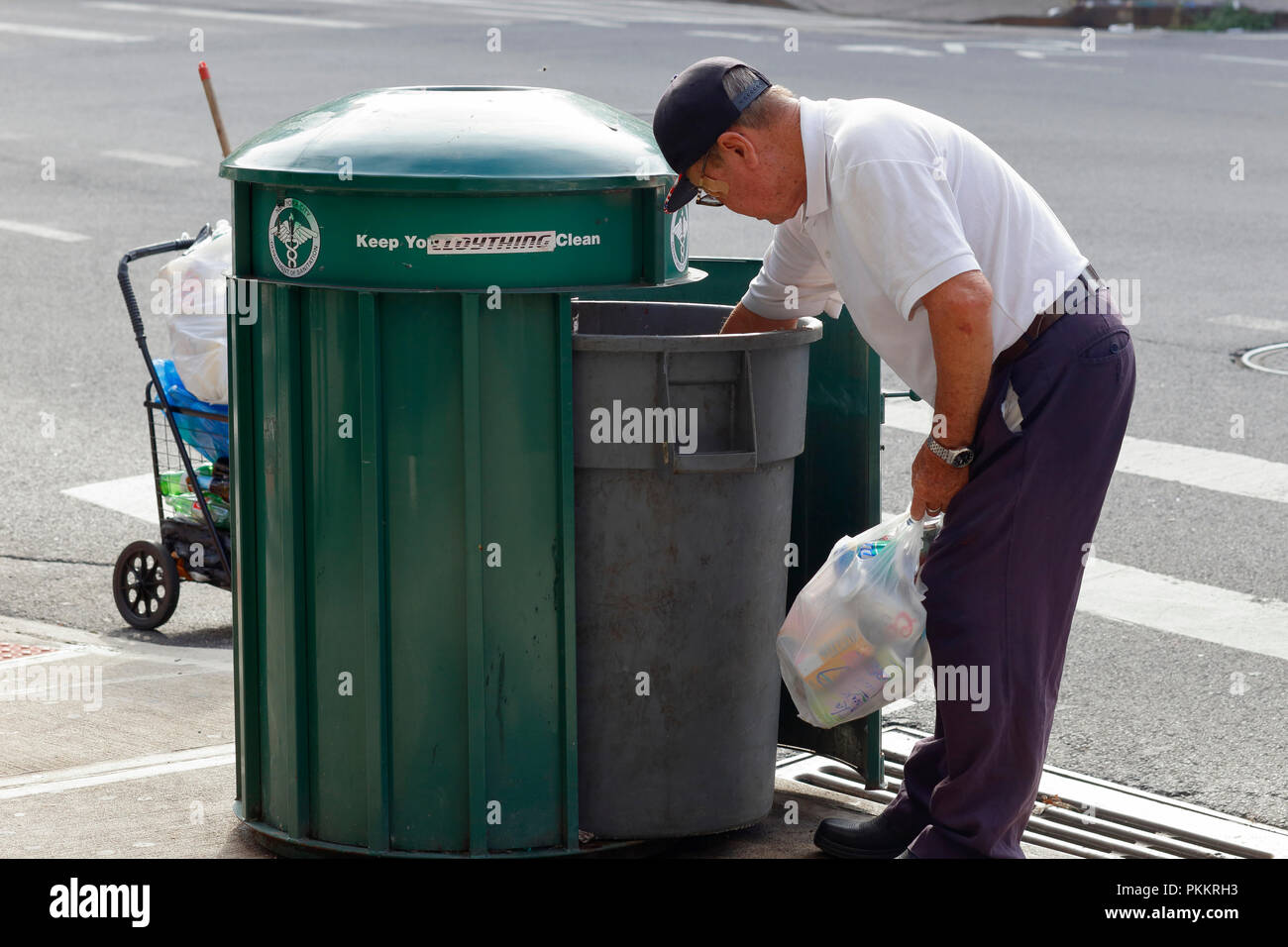An elderly Asian person reaches into a New York City trash bin for