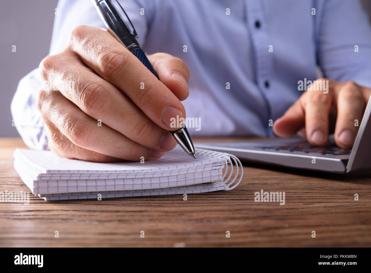 Person's Hand Writing On Black Notebook Using Pen Over Laptop Stock Photo