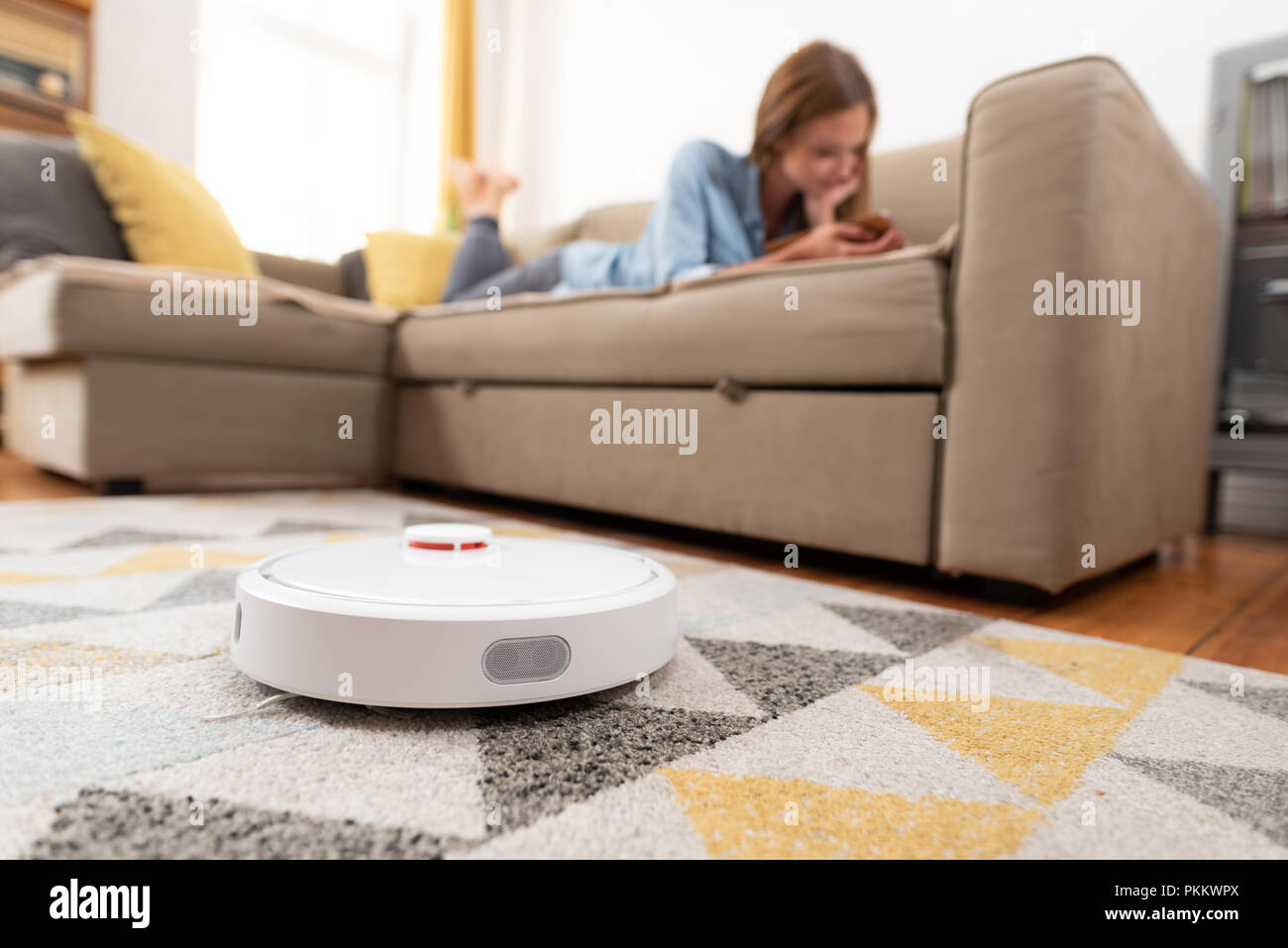 Robotic vacuum cleaner cleaning the room while woman relaxing on sofa. Woman controlling vacuum with remote control. Stock Photo