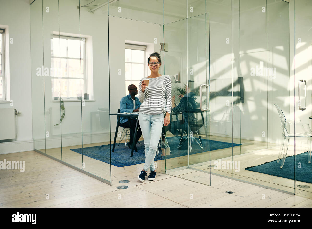 Young Asian businesswoman wearing glasses and smiling while drinking a coffee by the door of an office boardroom with coworkers in the background Stock Photo