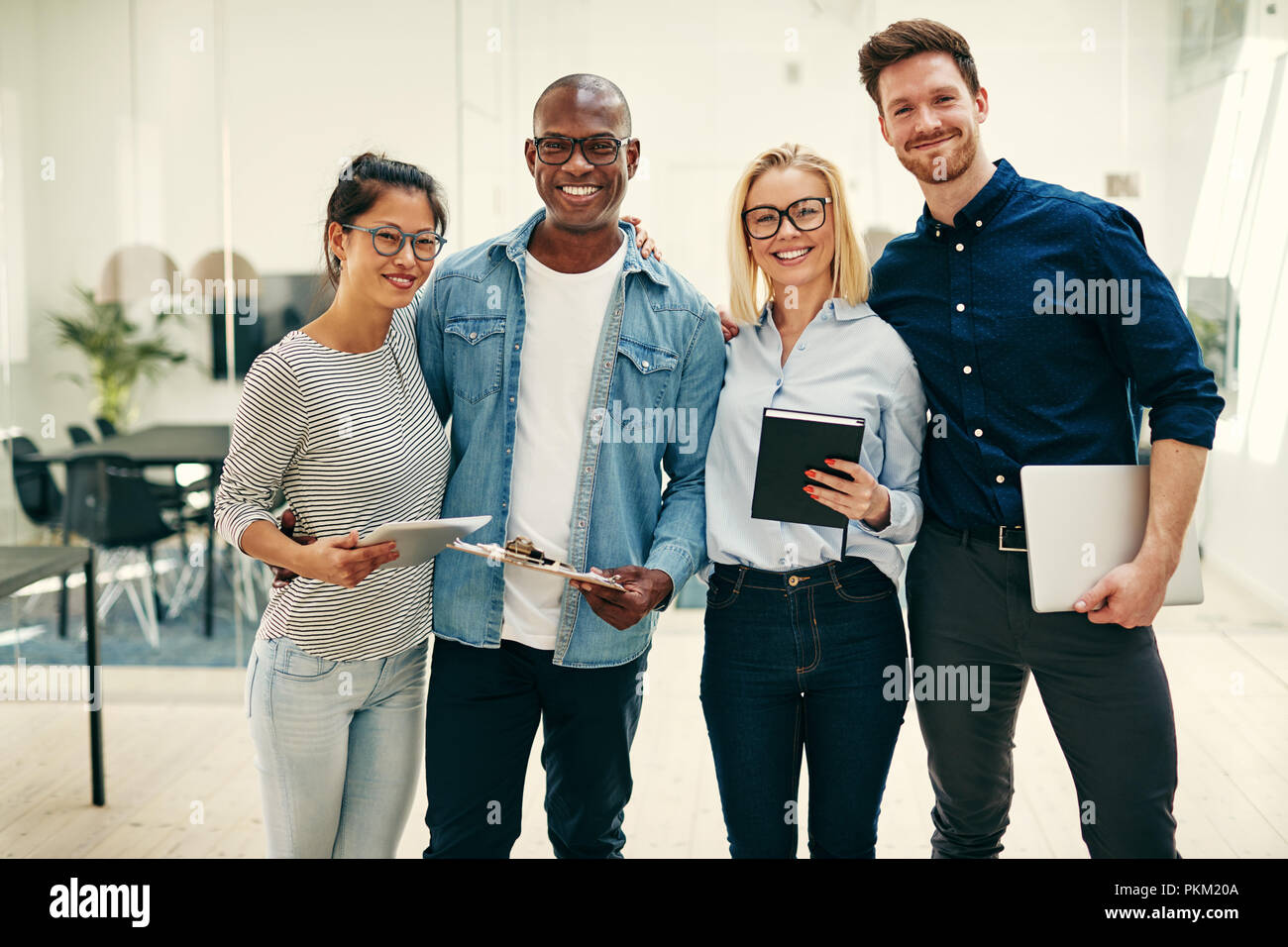 Diverse group of smiling young businesspeople standing together in a bright modern office ready for the business day Stock Photo