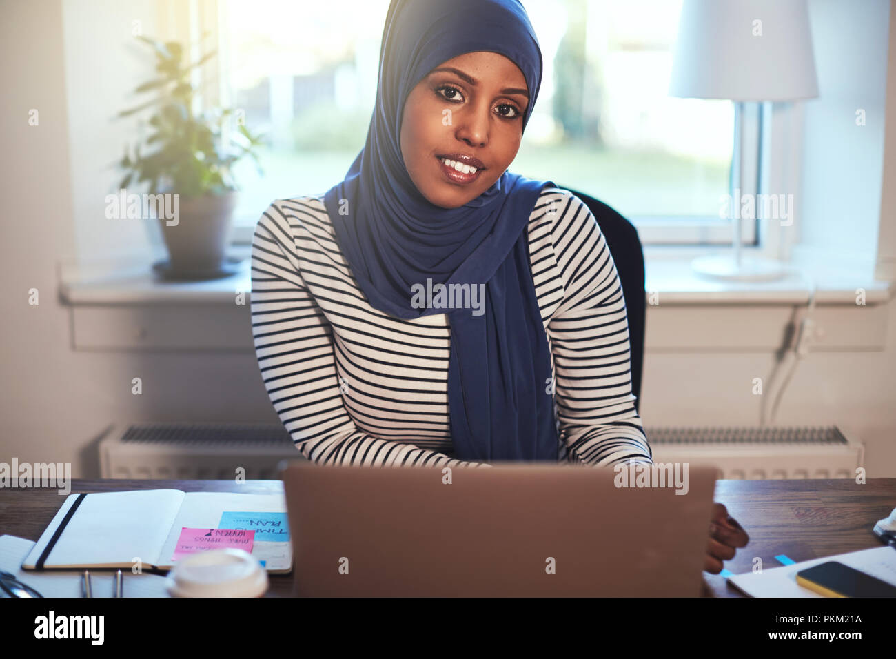 Smiling young Arabic female entrepreneur wearing a hijab sitting at a ...