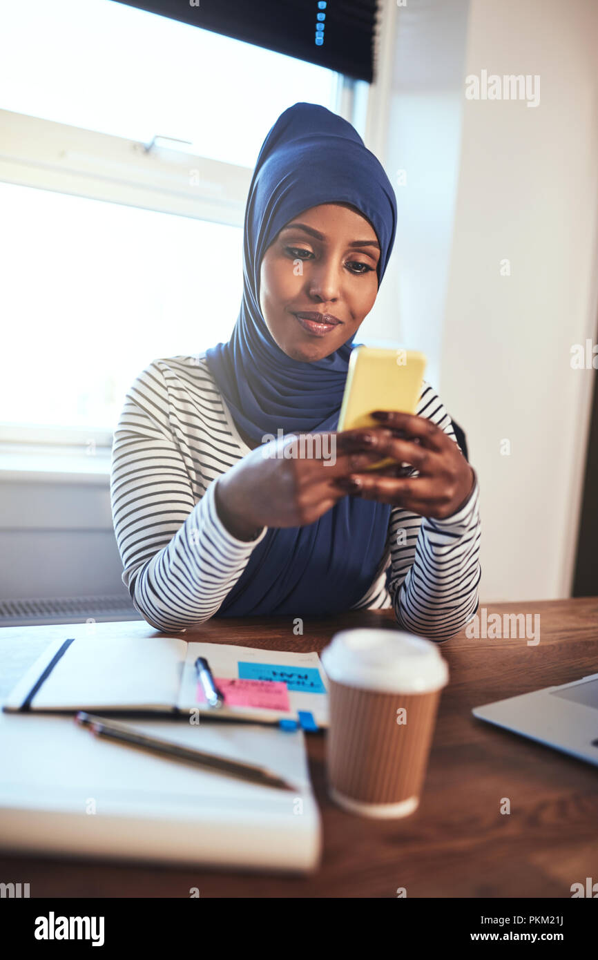 Smiling young Arabic female entrepreneur wearing a hijab sending a text ...