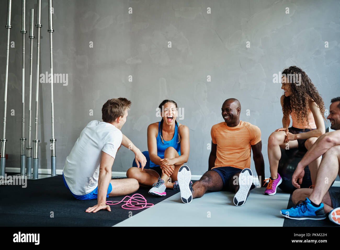 Friends in sportswear talking and laughing together while sitting on the floor of a gym after a workout Stock Photo